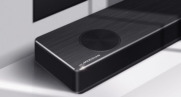 Κοντινό πλάνο της αριστερής πλευράς του LG Soundbar με το λογότυπο της Meridian στην κάτω αριστερή γωνία του προϊόντος. Η κάτω αριστερή πλευρά της τηλεόρασης είναι επίσης ορατή.