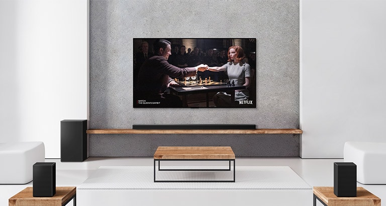 Ένα σετ με 2 πίσω ηχεία, ένα subwoofer και ένα soundbar και μια τηλεόραση σε ένα σαλόνι με λευκή απόχρωση. Στην οθόνη της τηλεόρασης φαίνεται ένας άντρας και μια γυναίκα που παίζουν σκάκι.