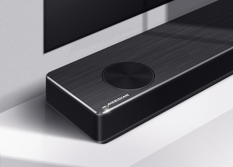 Κοντινό πλάνο της αριστερής πλευράς του LG Soundbar με το λογότυπο της Meridian στην κάτω αριστερή γωνία του προϊόντος. Η κάτω αριστερή πλευρά της τηλεόρασης είναι επίσης ορατή.