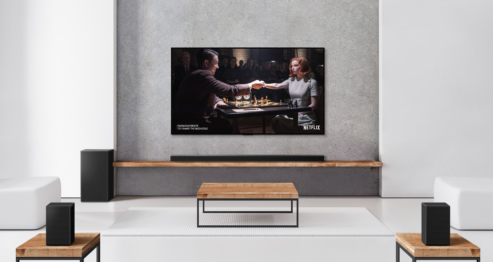 Ένα σετ με 2 πίσω ηχεία, ένα subwoofer και ένα soundbar και μια τηλεόραση σε ένα σαλόνι με λευκή απόχρωση. Στην οθόνη της τηλεόρασης φαίνεται ένας άντρας και μια γυναίκα που παίζουν σκάκι.