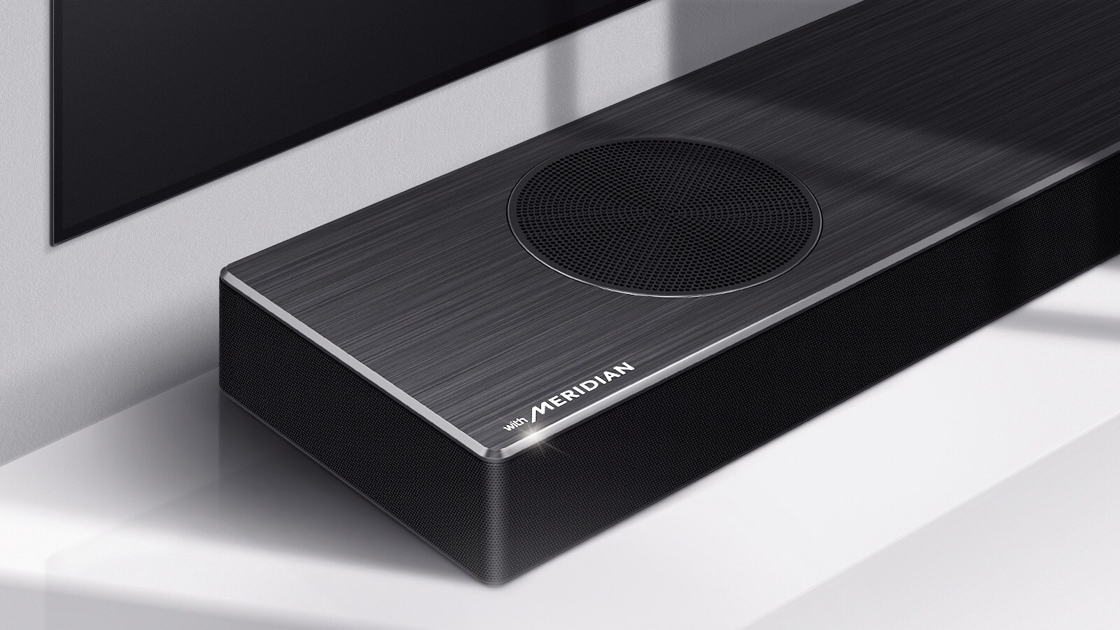 Κοντινό πλάνο της αριστερής πλευράς του LG Soundbar με το λογότυπο της Meridian στην κάτω αριστερή γωνία του προϊόντος. Η κάτω αριστερή πλευρά της τηλεόρασης είναι επίσης ορατή.