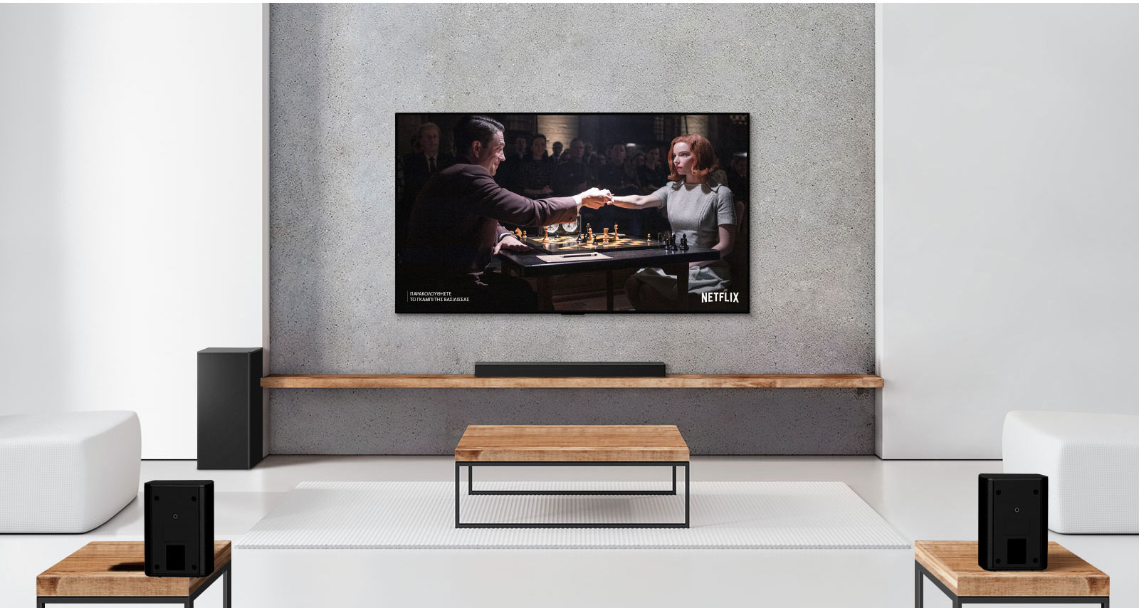 Ένα σετ με 2 πίσω ηχεία, ένα subwoofer και ένα soundbar και μια τηλεόραση σε ένα σαλόνι με λευκή απόχρωση. Στην οθόνη της τηλεόρασης φαίνεται ένας άντρας και μια γυναίκα που παίζουν σκάκι. 