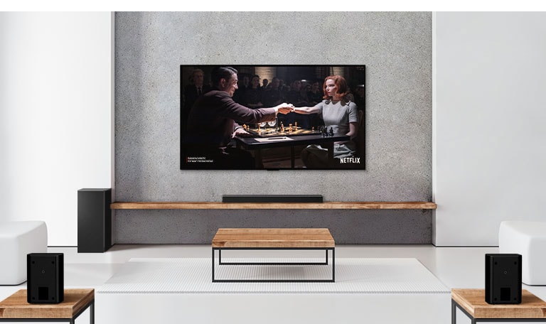 Ένα σετ με 2 πίσω ηχεία, ένα subwoofer και ένα soundbar και μια τηλεόραση σε ένα σαλόνι με λευκή απόχρωση. Στην οθόνη της τηλεόρασης φαίνεται ένας άντρας και μια γυναίκα που παίζουν σκάκι. 