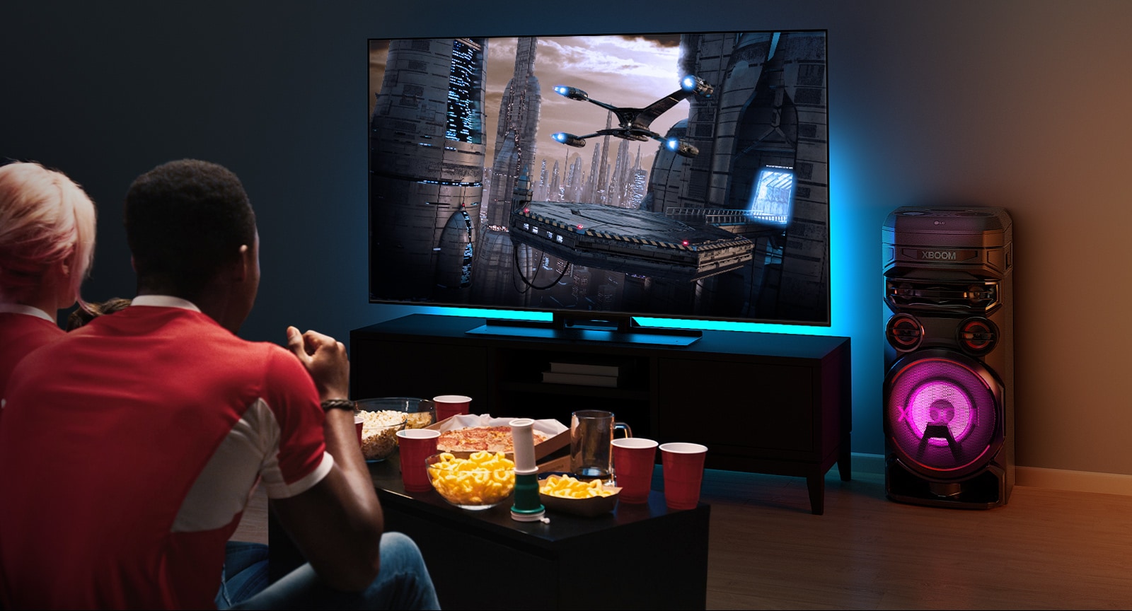 Άτομα που παρακολουθούν τηλεόραση σε έναν τοίχο με ένα LG XBOOM RNC7 στα δεξιά. Η τηλεόραση προβάλλει μια ταινία. 
