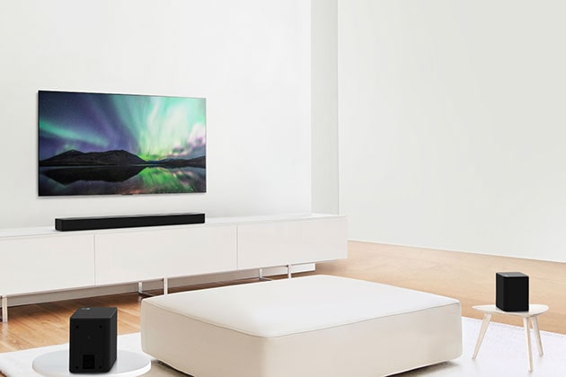 Τηλεόραση και Sound Bar σε σαλόνι με λευκή διακόσμηση, με λευκό καναπέ στο κέντρο. Εκατέρωθεν του καναπέ υπάρχουν δύο ηχεία.