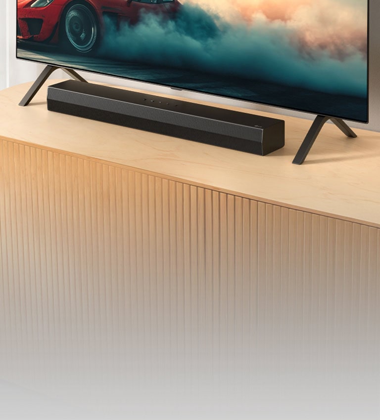 Το LG Soundbar S20A τοποθετείται σε μια ξύλινη κονσόλα τηλεόρασης, κάτω από μια τηλεόραση και ανάμεσα στις βάσεις της τηλεόρασης. Στην τηλεόραση εμφανίζεται ένα κόκκινο αυτοκίνητο με καπνό.