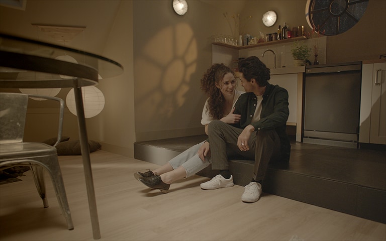 Μπροστά από το πλυντήριο πιάτων, ένα ζευγάρι κάθεται δίπλα-δίπλα χαμογελώντας ο ένας στον άλλον. Αυτή η εικόνα διαδέχεται την άλλη εικόνα. Φαίνεται το εσωτερικό του πλυντηρίου πιάτων, όπου πλένονται τα πιάτα.