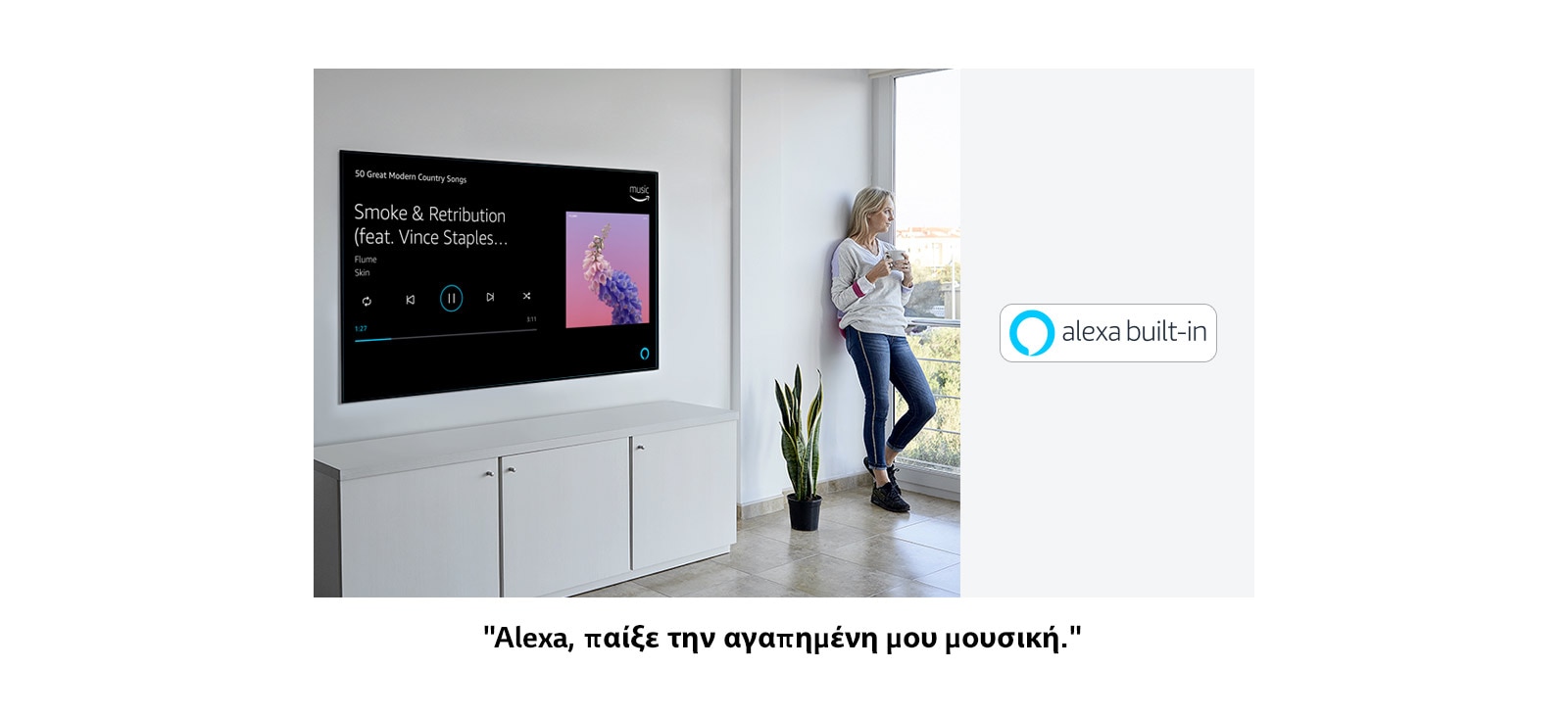 Μια γυναίκα που γέρνει στον τοίχο δίπλα στην τηλεόραση, κοιτάζοντας έξω από το παράθυρο και ζητώντας από την Alexa να παίξει την αγαπημένη της μουσική.