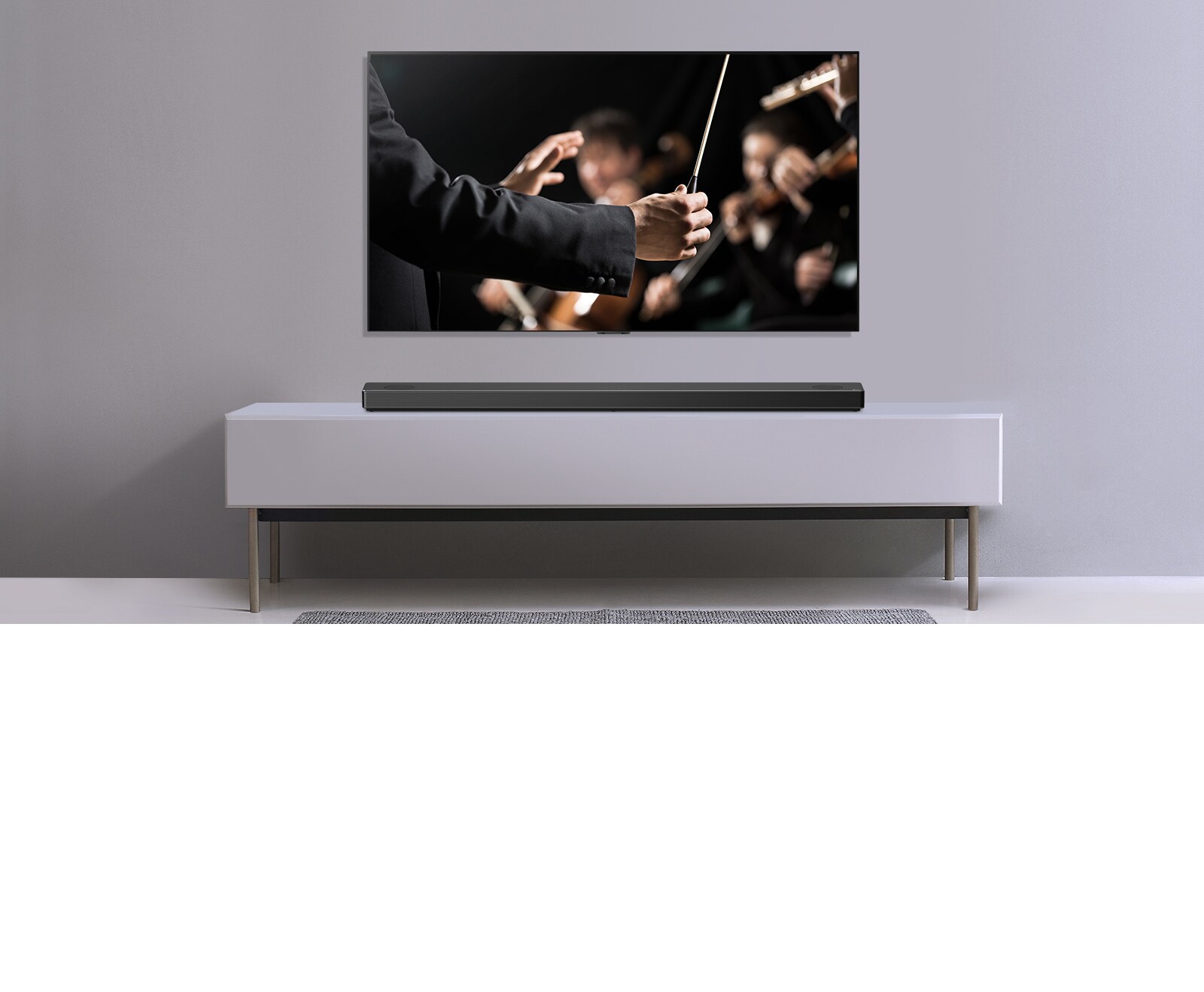 Μια TV σε γκρίζο τοίχο και από κάτω σε ένα γκρίζο ράφι - ένα LG Sound Bar. Στην τηλεόραση ένας μαέστρος διευθύνει ορχήστρα.