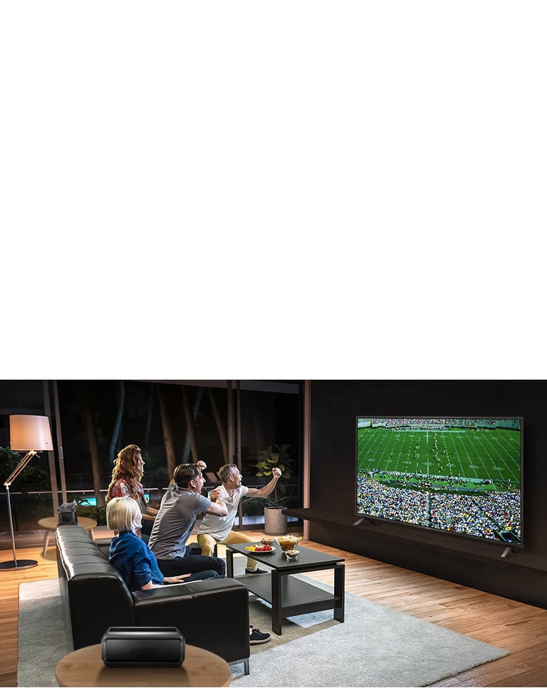 Μια παρέα παρακολουθεί αθλητικά παιχνίδια στην τηλεόραση στο σαλόνι, με τα ηχεία Bluetooth πίσω τους.
