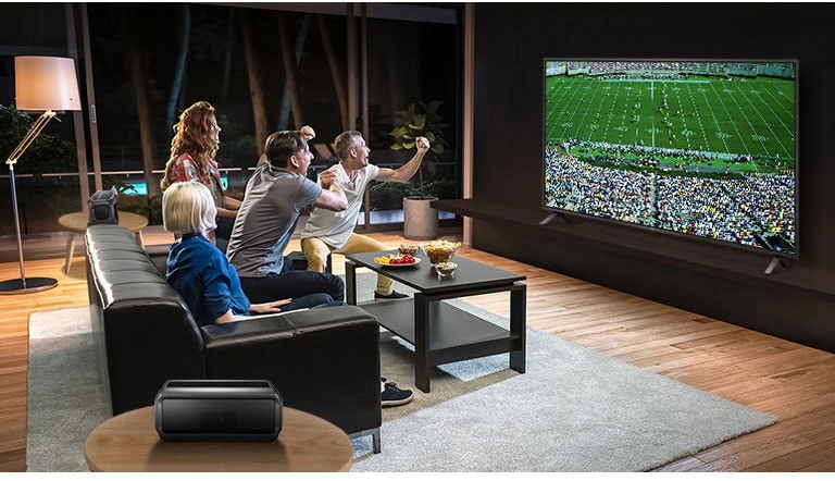 Μια παρέα παρακολουθεί αθλητικά παιχνίδια στην τηλεόραση στο σαλόνι, με τα ηχεία Bluetooth πίσω τους.