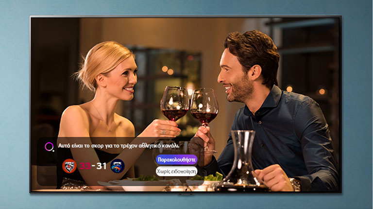 Μια οθόνη τηλεόρασης που απεικονίζει έναν άνδρα και μια γυναίκα που τσουγκρίζουν τα ποτήρια τους, ενώ εμφανίζονται αθλητικές ειδοποιήσεις