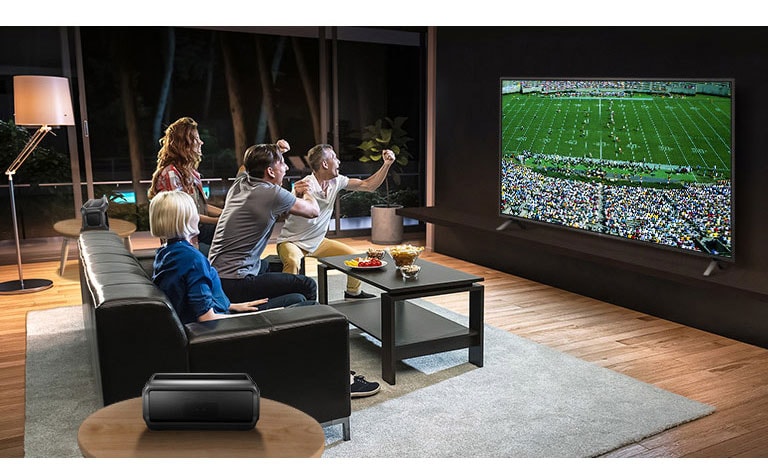 Μια παρέα παρακολουθεί αθλητικά παιχνίδια στην τηλεόραση στο σαλόνι, με τα ηχεία Bluetooth πίσω τους.