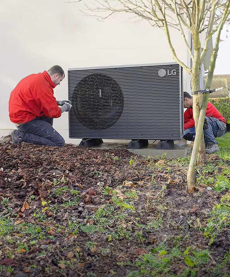 Δύο τεχνικοί εγκατάστασης με κόκκινες φόρμες εγκαθιστούν την αντλία θερμότητας αέρα/νερού THERMA V R290 Monobloc της LG στην αυλή ενός σπιτιού.