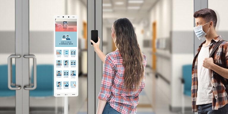 Ένας χρήστης σαρώνει έναν κωδικό QR με το κινητό του τηλέφωνο σε ένα τερματικό θερμικής ανίχνευσης 29" της LG που είναι εγκατεστημένο μπροστά από την είσοδο.