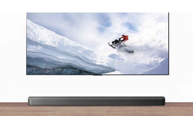 Η τηλεόραση & το Soundbar φαίνονται από εμπρός. Η τηλεόραση δείχνει έναν άντρα επάνω σε ένα όχημα χιονιού σε χιονισμένα βουνά. Το λογότυπο του HDMI 2.1 βρίσκεται κάτω από την τηλεόραση.
