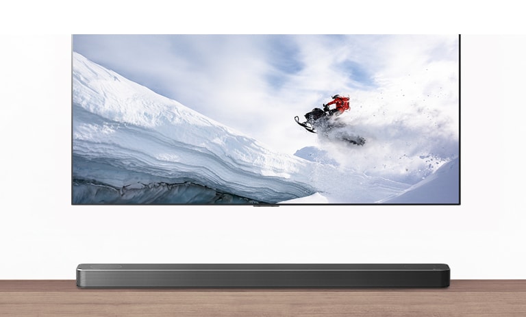 Η τηλεόραση & το Soundbar φαίνονται από εμπρός. Η τηλεόραση δείχνει έναν άντρα επάνω σε ένα όχημα χιονιού σε χιονισμένα βουνά. Το λογότυπο του HDMI 2.1 βρίσκεται κάτω από την τηλεόραση.