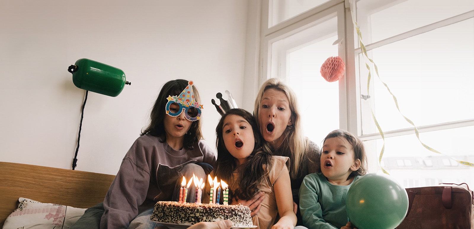 Μια εικόνα δύο γυναικών και ενός κοριτσιού που φοράει ένα καπελάκι γενεθλίων και φυσάει τα κεριά σε μια τούρτα.