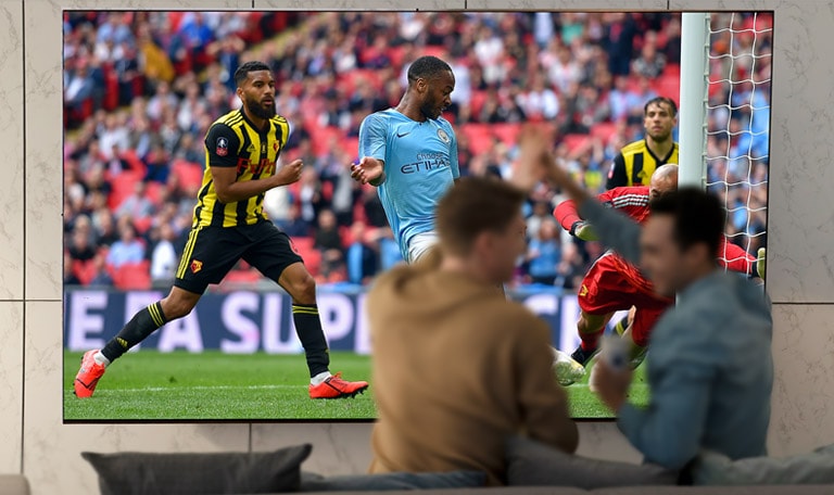 Μια παρέα παρακολουθεί έναν αγώνα σε τηλεόραση μεγάλων διαστάσεων σε ένα σαλόνι σπιτιού.