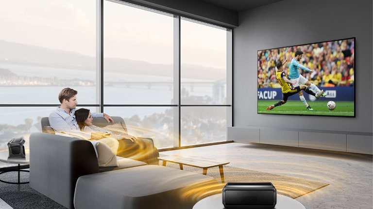 Μια παρέα ανδρών και γυναικών παρακολουθεί έναν αγώνα στην τηλεόραση ενός σαλονιού, με τα ηχεία Bluetooth πίσω τους.