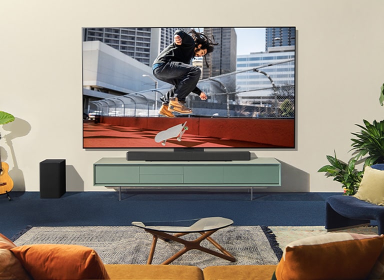 Dvije žene sjede na sofi ispred LG televizora montiranog na zidu u raznobojnom dnevnom boravku, dok jedna žena usmjerava daljinsku upravljač prema zaslonu televizora. 