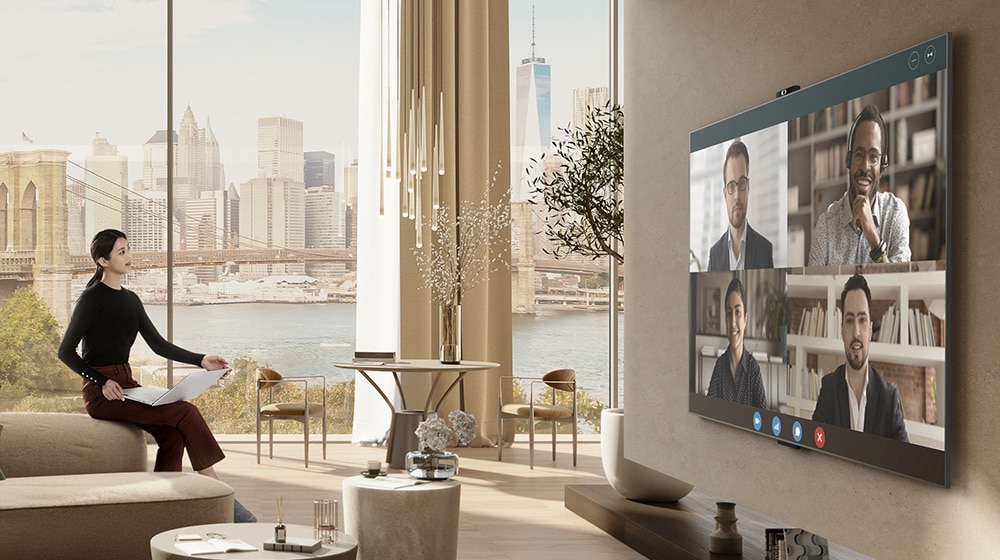 Žena sjedi na rubu kauča, drži prijenosno računalo i gleda televiziju. Na televizoru na velikom zaslonu možete vidjeti četiri osobe i videokonferenciju.