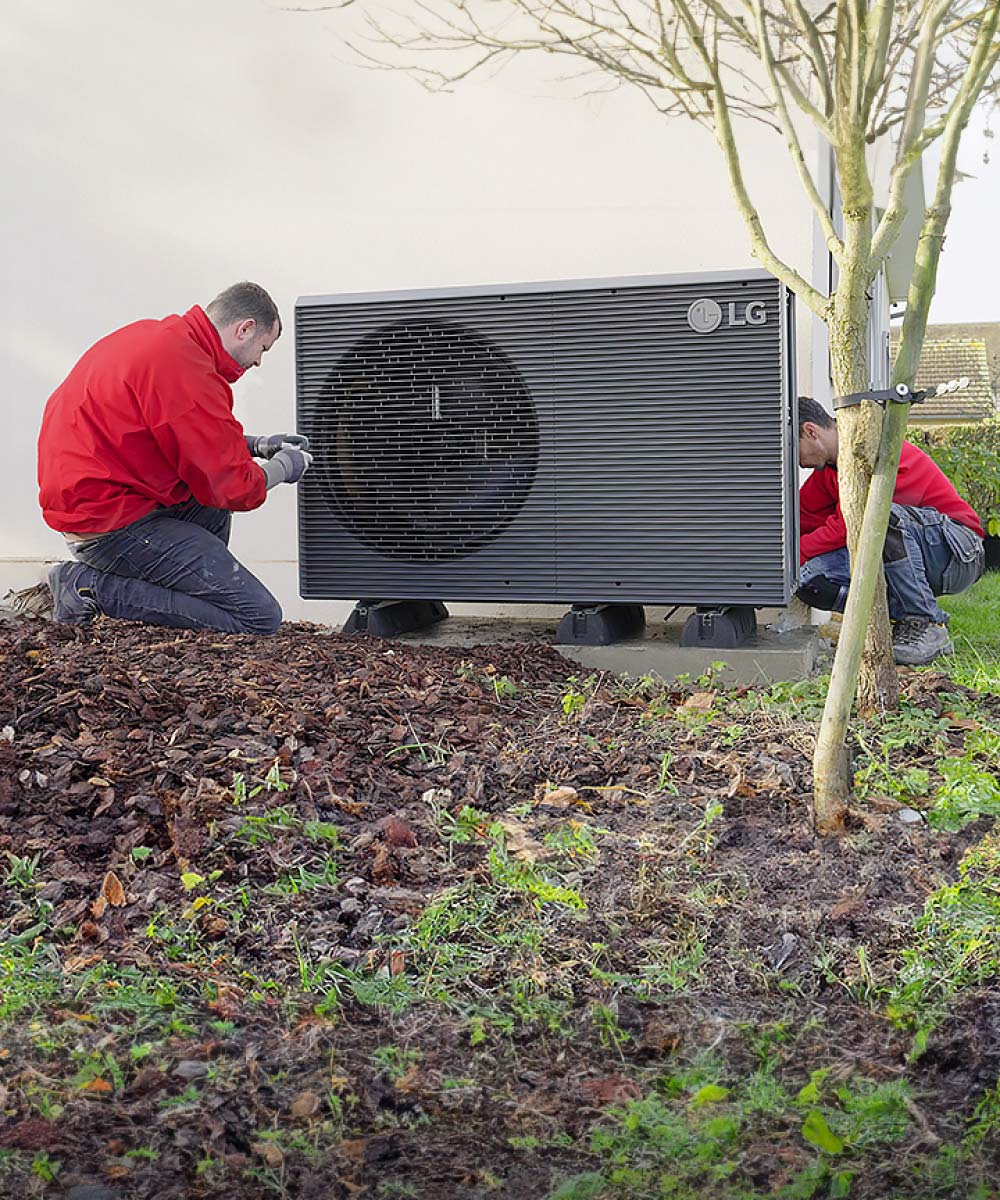 Dva instalatera u crvenim radnim uniformama ugrađuju LG zračno-vodenu dizalicu topline THERMA V R290 Monobloc u dvorištu kuće.