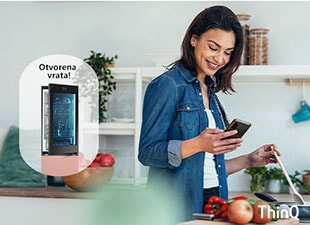 A képen egy nő főzés közben értesítést kap a mobiltelefonjára az LG ThinQ applikációból, a nyitva felejtett hűtő ajtó miatt.