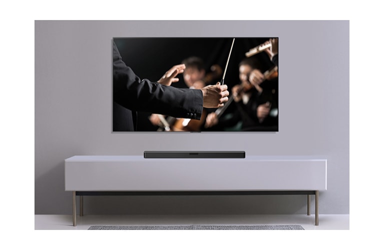 Na sivom zidu nalazi se televizor, a ispod njega na sivoj polici zvučnik LG Sound Bar. Na televizoru je prikazan dirigent koji dirigira orkestrom.