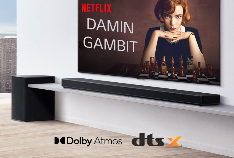 Televizor se nalazi na zidu. Plakat televizijske emisije je na zaslonu televizora. Zvučnik LG Soundbar nalazi se neposredno ispod televizora na bijeloj polici, a neposredno pored njega vidi se niskofrekvencijski zvučnik. Logotipi Dolby Atmos i DTS:X prikazani pri dnu slike u sredini.