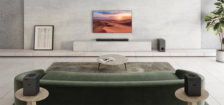 Televizor visi na bijelom stupu u sredini dnevnog boravka. Ispod je LG Sound Bar smješten na bijeloj polici. Na podu ispod zvučnika nalazi se bežični niskofrekvencijski zvučnik. Iza zelene sofe dva stražnja zvučnika smještena su na stoliću s lijeve i desne strane. Oznaka NOVO prikazana je u gornjem lijevom kutu.