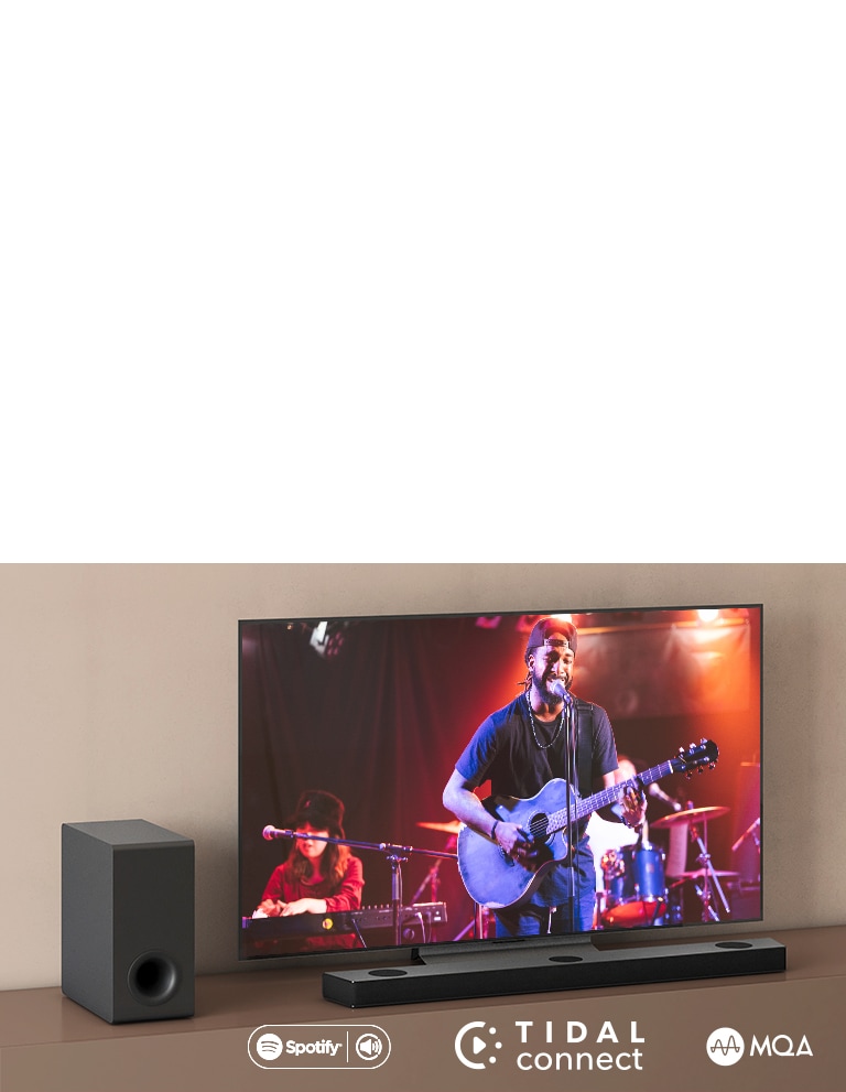LG televizor nalazi se na smeđoj polici, LG Sound Bar S80QY nalazi se ispred televizora. Niskofrekvencijski zvučnik smješten je s lijeve strane televizora. Na televizoru se prikazuje scena koncerata. Oznaka NOVO prikazana je u gornjem lijevom kutu.