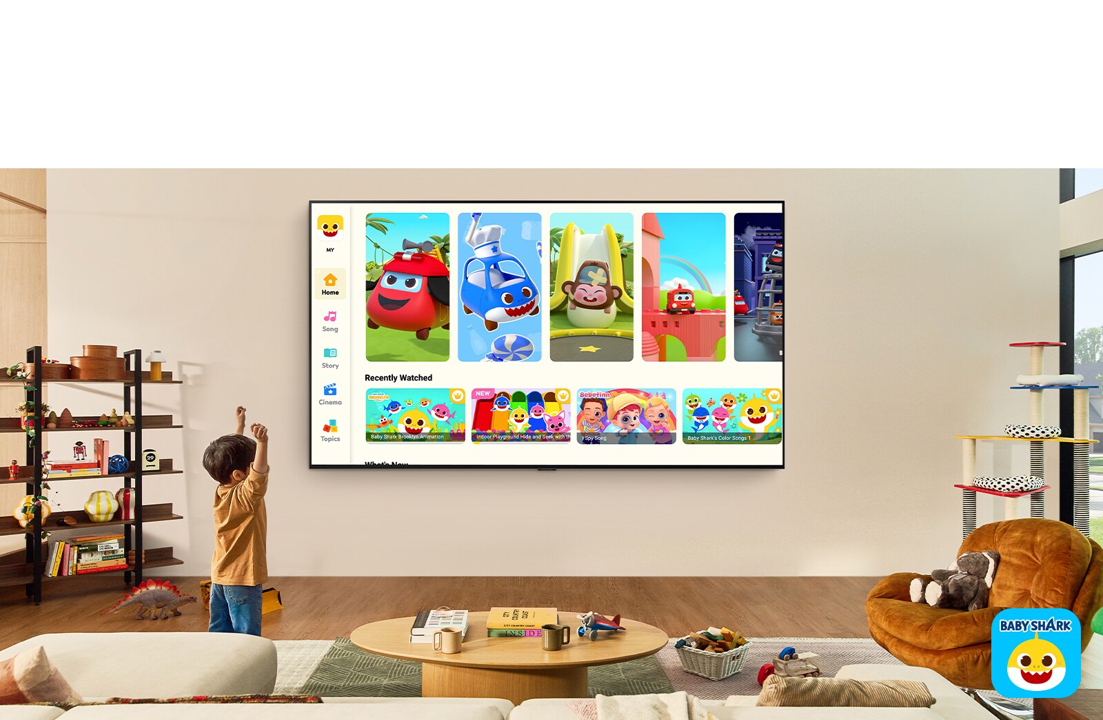 ילד קטן צופה ב-Pinkfong על LG TV התלויה על הקיר בחלל מגורים עם צעצועים לילדים. 