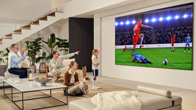 משפחה הנמצאת בסלון שלהם. יש להם LG TV תלויה על הקיר עם מסך גדול במיוחד שגדול כמעט כמו הקיר כולו. הם צופים במשחק כדורגל מרגש על המסך הגדול והסוחף.