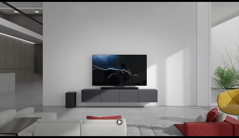 מקרן הקול ניצב על מזנון אפור בסלון יחד עם הטלוויזיה בסלון. סאב וופר אלחוטי שחור ניצב על הרצפה בצד השמאלי ואור השמש בוקע מהצד הימני של התמונה. ספה ארוכה בגווני לבן וורוד ניצבת מול הטלוויזיה ומקרן הקול.