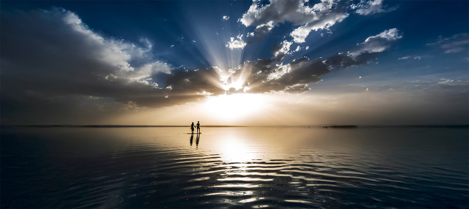 השמש הזורחת מעל האופק מוסתרת בחלקה על ידי עננים, שולחת קרני אור אל פני השמיים הכהים, בעוד שהשתקפותה על פני המים יוצרת ניגוד עדין מול החשכה שסביבה.