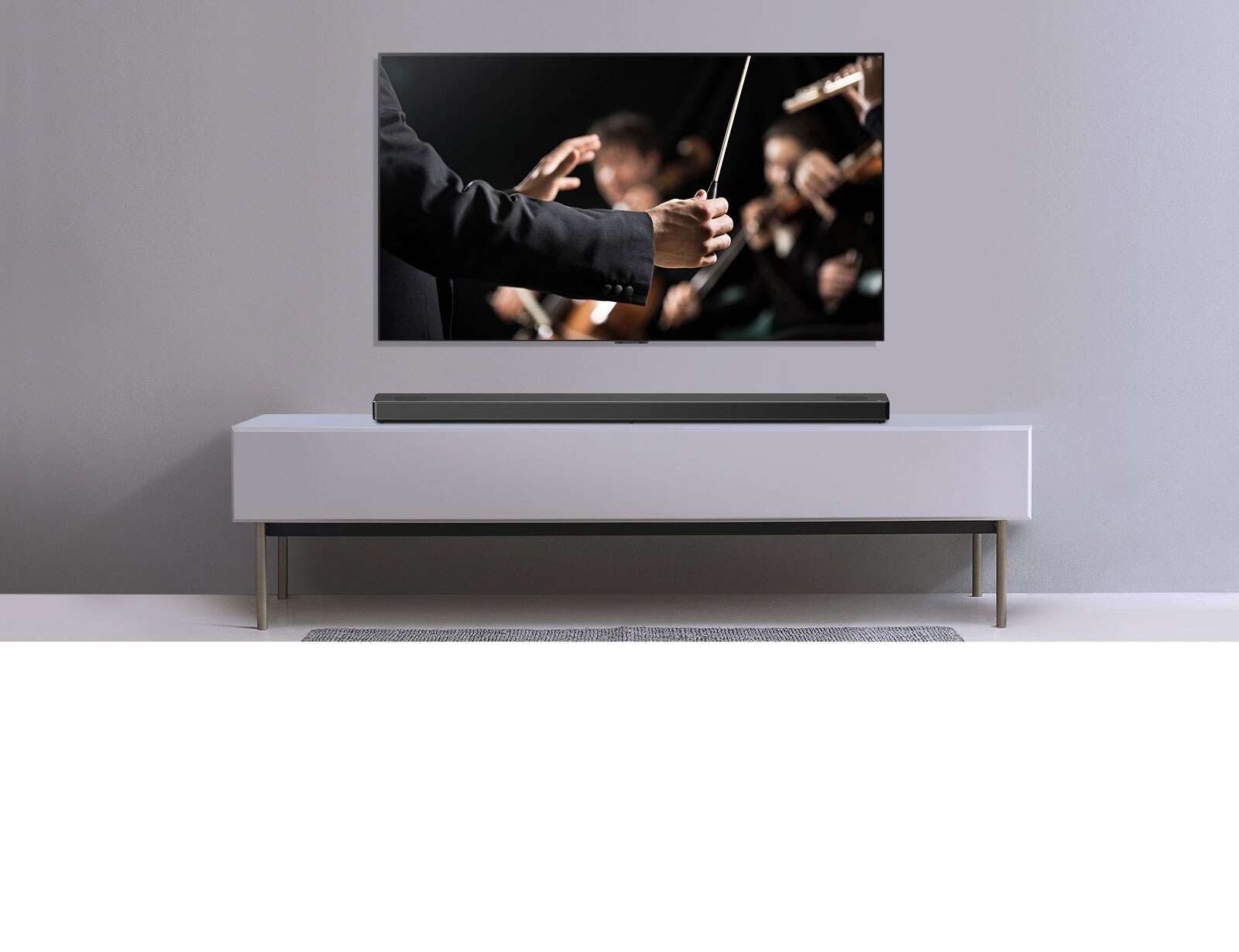טלוויזיה מוצגת על קיר אפור והסאונד-בר מבית LG מונח מתחתיה על מזנון אפור. טלוויזיה מציגה מנצח תזמורת.