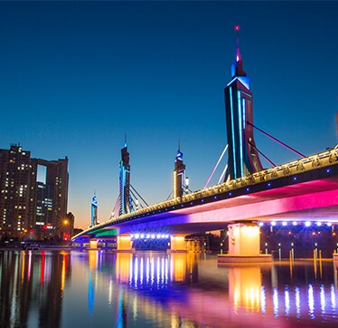 מבט לילי של גשר מודרני המואר באור בהיר על רקע גורדי שחקים, עם תאורה חיה המשתקפת במים, המדגימה ניגודיות גבוהה ועושר בפרטי צבע.