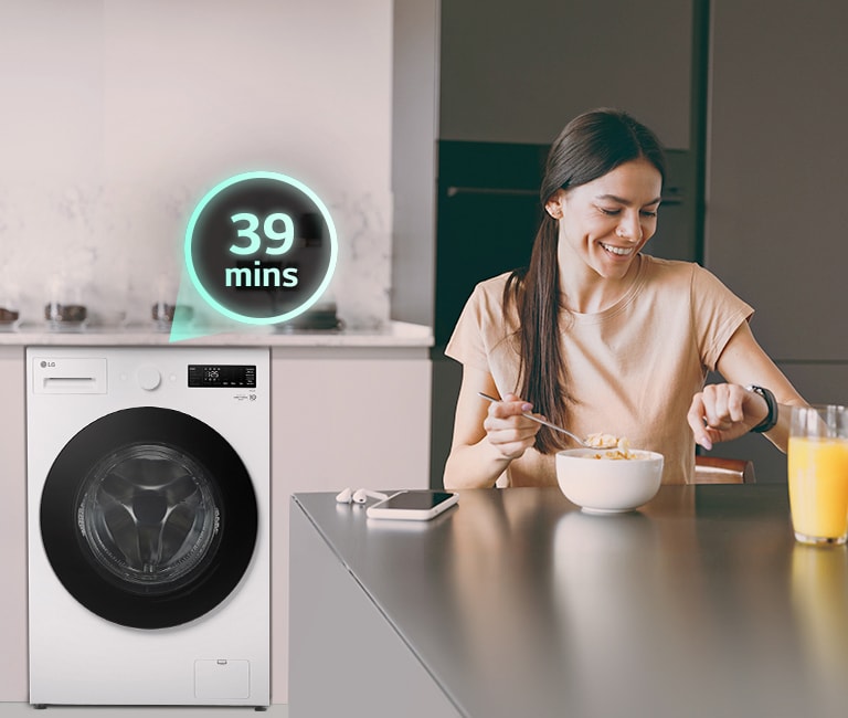 אישה יושבת בנחת ליד השולחן, מביטה בשעונה, כשברקע מכונת הכביסה פועלת