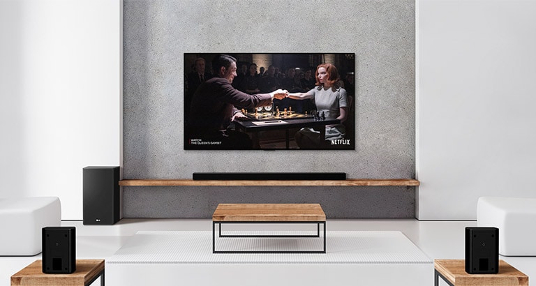 مجموعه‌ای از دو بلندگوی عقب، ساب ووفر، ساندبار و تلویزیون در یک اتاق نشیمن سفید جای گرفته‌اند. زن و مردی در حال بازی شطرنج در صفحه تلویزیون هستند.