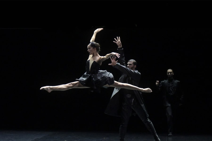 Tutti in costumi neri, una ballerina fa la spaccata a mezz’aria mentre altri due ballerini sono dietro di lei.