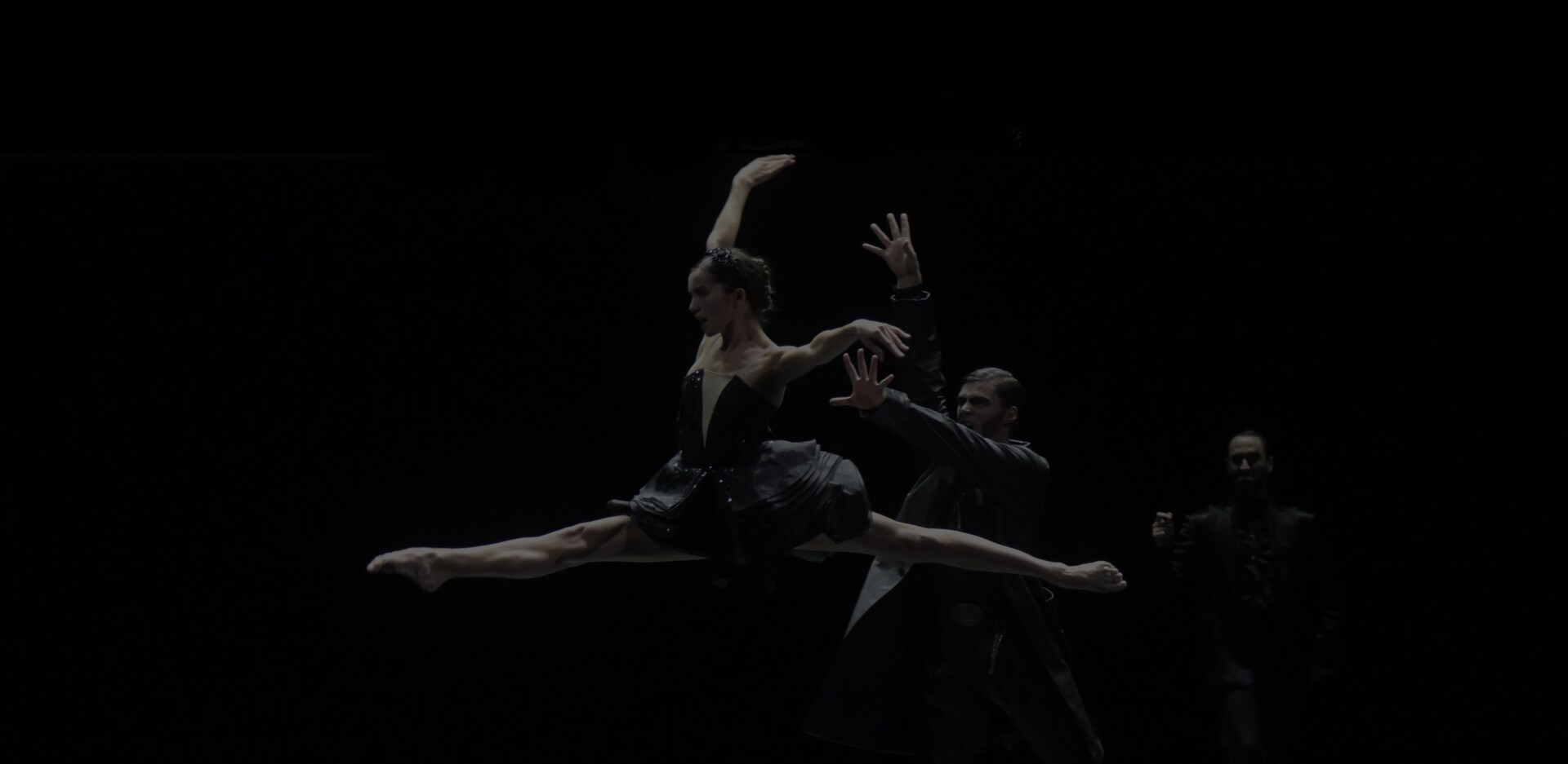 A female dancer is captured doing the splits mid-air, accompanied by a male dancer standing towards her. The two are clad in black costumes.