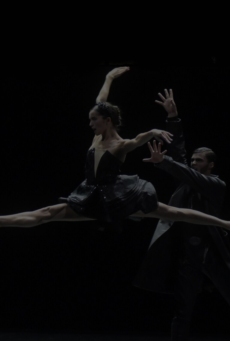 A female dancer is captured doing the splits mid-air, accompanied by a male dancer standing towards her. The two are clad in black costumes.