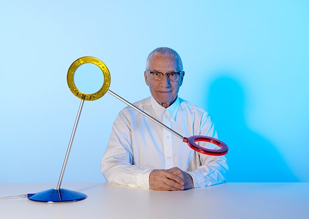 Alessandro Mendini intento a guardare diritto con la sua scultura artistica accanto a sé.