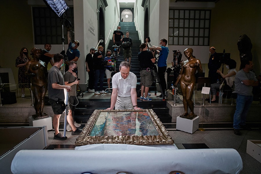 Uomo con lo sguardo abbassato su un'opera d'arte incorniciata mentre sul retro un team cinematografico predispone un set.