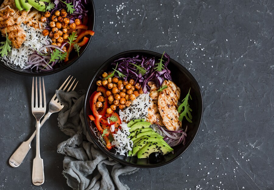 Due ciotole nere con abbondante insalata di pollo, ceci e avocado su una superficie di pietra nera con due forchette incrociate