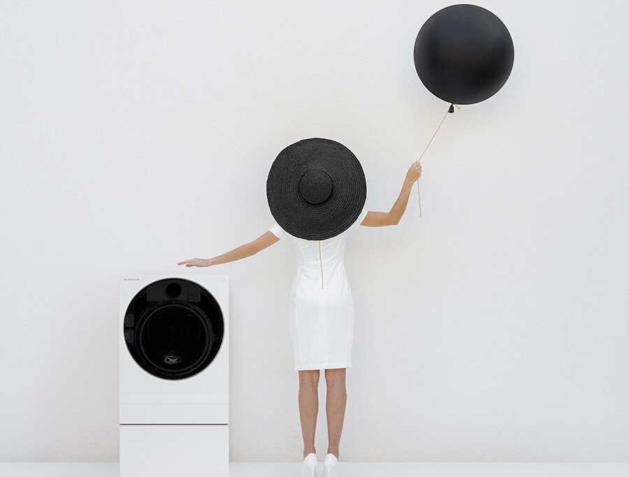 una lavatrice lg signature è posta sulla sinistra di uno spazio completamente bianco con al centro anna, in piedi con un vestito bianco, un cappello nero e un palloncino nero nella mano destra
