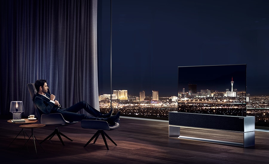 Un uomo guarda la TV arrotolabile LG SIGNATURE rivolto verso la veduta notturna della città di Las Vegas nel proprio salotto.