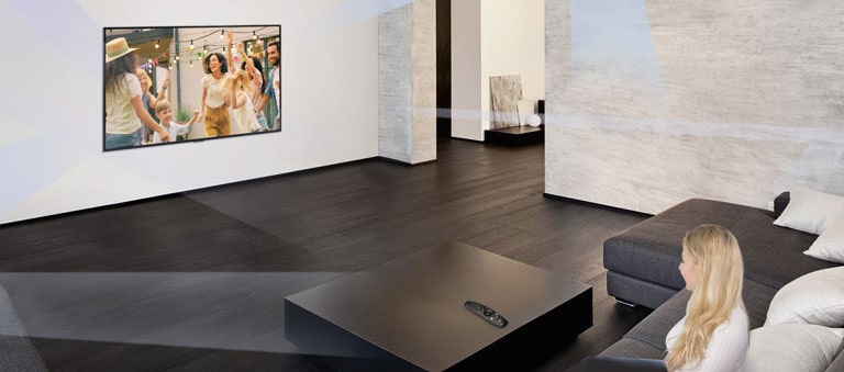 امرأة تجلس في غرفة معيشة رحبة تتميز بديكور داخلي بسيط وتشاهد أشخاصًا يرقصون على شاشة التلفزيون