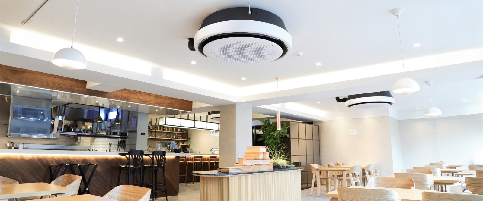 Wood and white tone dining area with tables, chairs, and kitchen displayed. LG Round Cassette mounted on the ceiling.